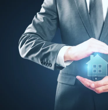 Close-up of a businessman in a grey suit protecting a glowing house icon between his hands, symbolizing real estate asset protection and security.